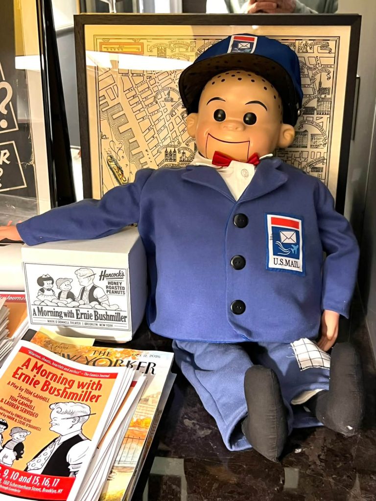 A ventriloquist dummy wearing a U.S. Mail postal worker uniform, seated next to a box of peanuts and playbills for A Morning with Ernie Bushmiller, identified as 851924317_1049027581495192_3933971952008591539_n.